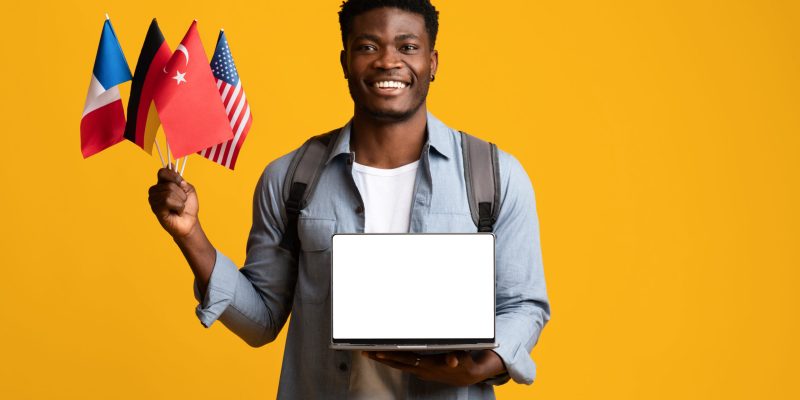 Happy black guy student wirh backpack holding various flags and laptop with empty screen, learning foreign languages on Internet, using website or online courses, mockup, yellow studio background