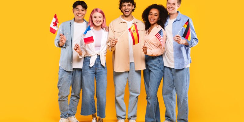 Happy diverse students of language school, teens of different nationalities holding little flags, posing on yellow background, studio shot, full length
