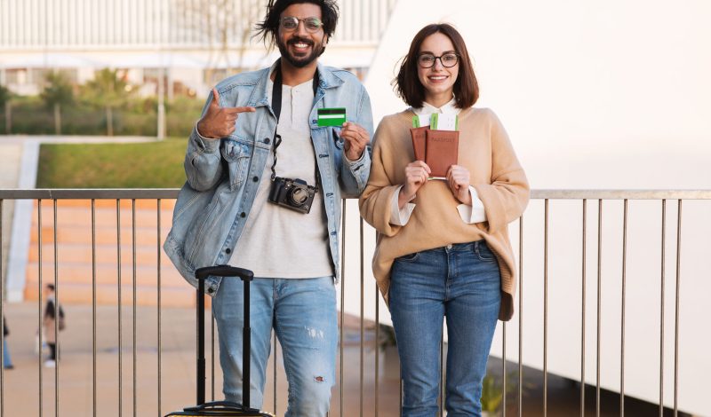 Positive millennial indian male and caucasian lady tourists with suitcase show passports, tickets and credit card in airport. Vacation and trip together, booking tour, cashback