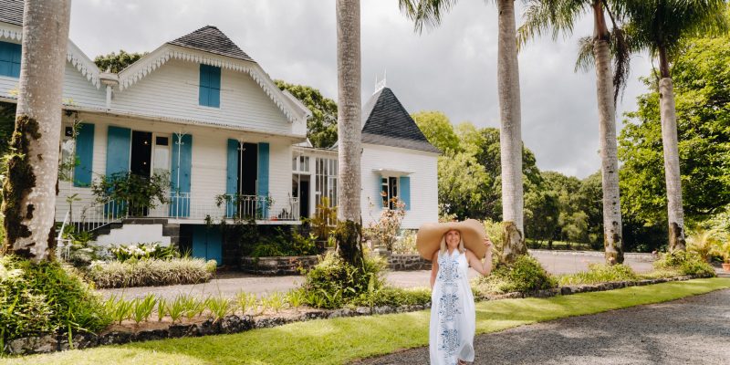 the woman in the big hat is smiling. a beautiful girl in a big hat and white dress smiles outside an old colonial building on the island of Mauritius.